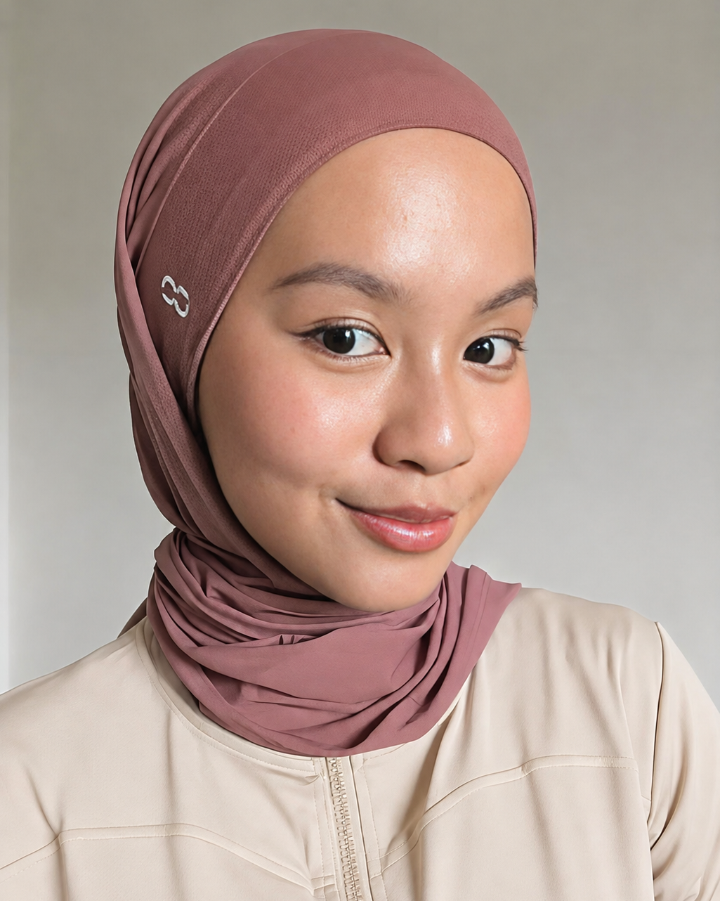 A woman wearing a dusty rose hijab and a beige zip-up jacket, photographed against a neutral light gray background.